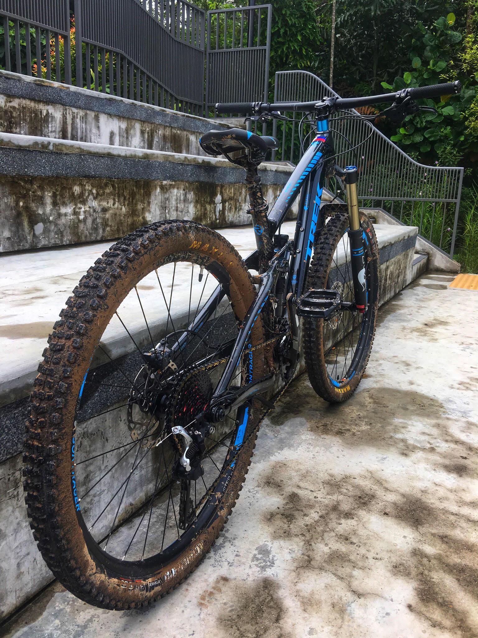 Trek Remedy 8: A mountain bike with a blue and black frame is leaning against a concrete surface, showcasing muddy tires and a dirty rear. The background features a set of steps and greenery, suggesting an outdoor environment possibly used for biking trails.