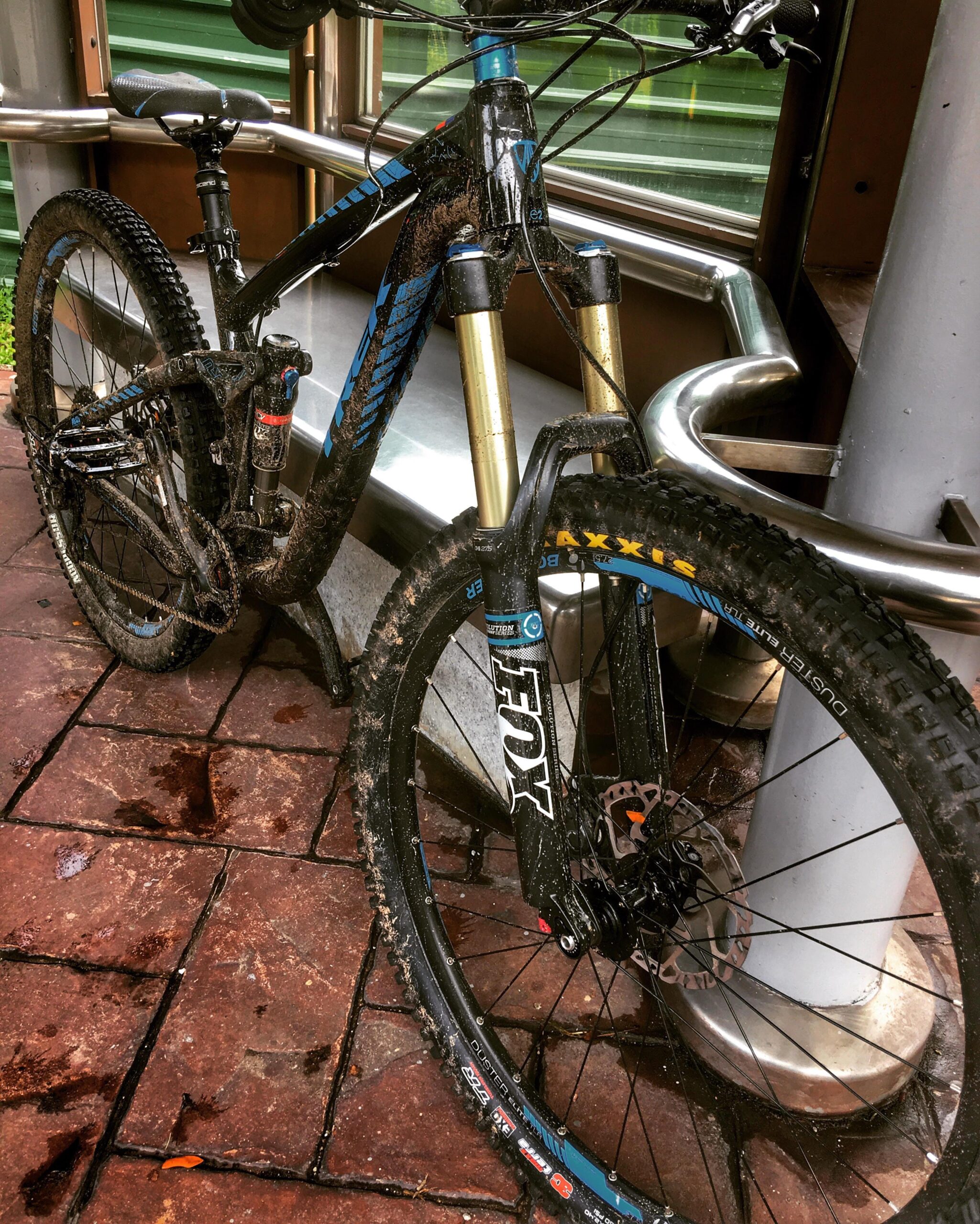 Trek Remedy 8: A muddy mountain bike resting on a paved surface, with visible dirt on the frame and tires. The bike features a black and blue design and is positioned near a metallic railing.