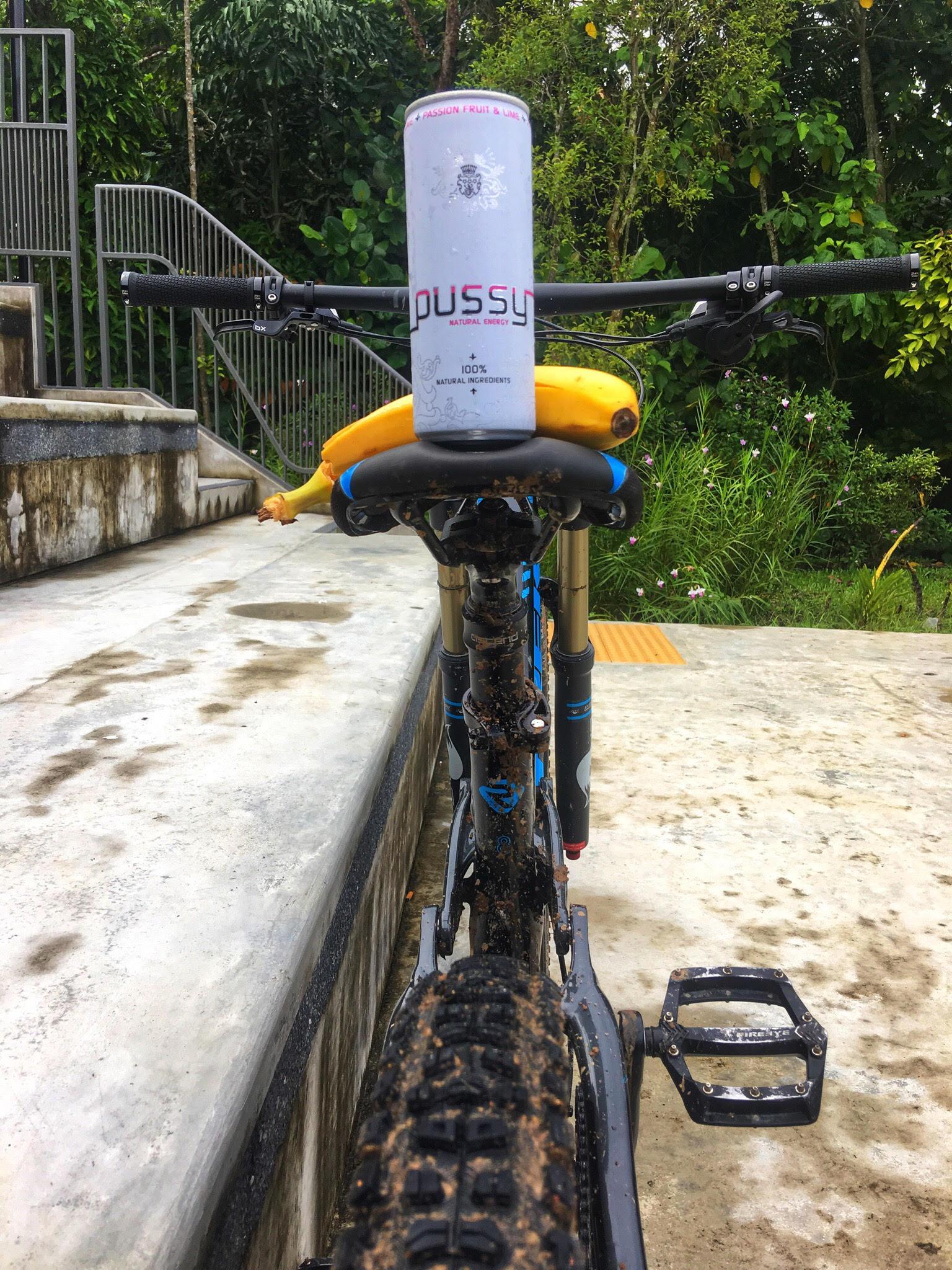 Trek Remedy 8: A mountain bike parked on a wet surface, displaying a can of energy drink on the seat and a banana resting beside it. The bike is muddy, indicating recent use, with greenery in the background.