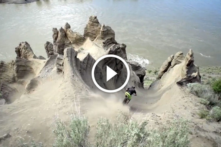 A mountain biker descending a steep, sandy slope with rocky formations in the background, overlooking a river. Dust is kicked up as the biker navigates the terrain, surrounded by greenery and rugged landscape.