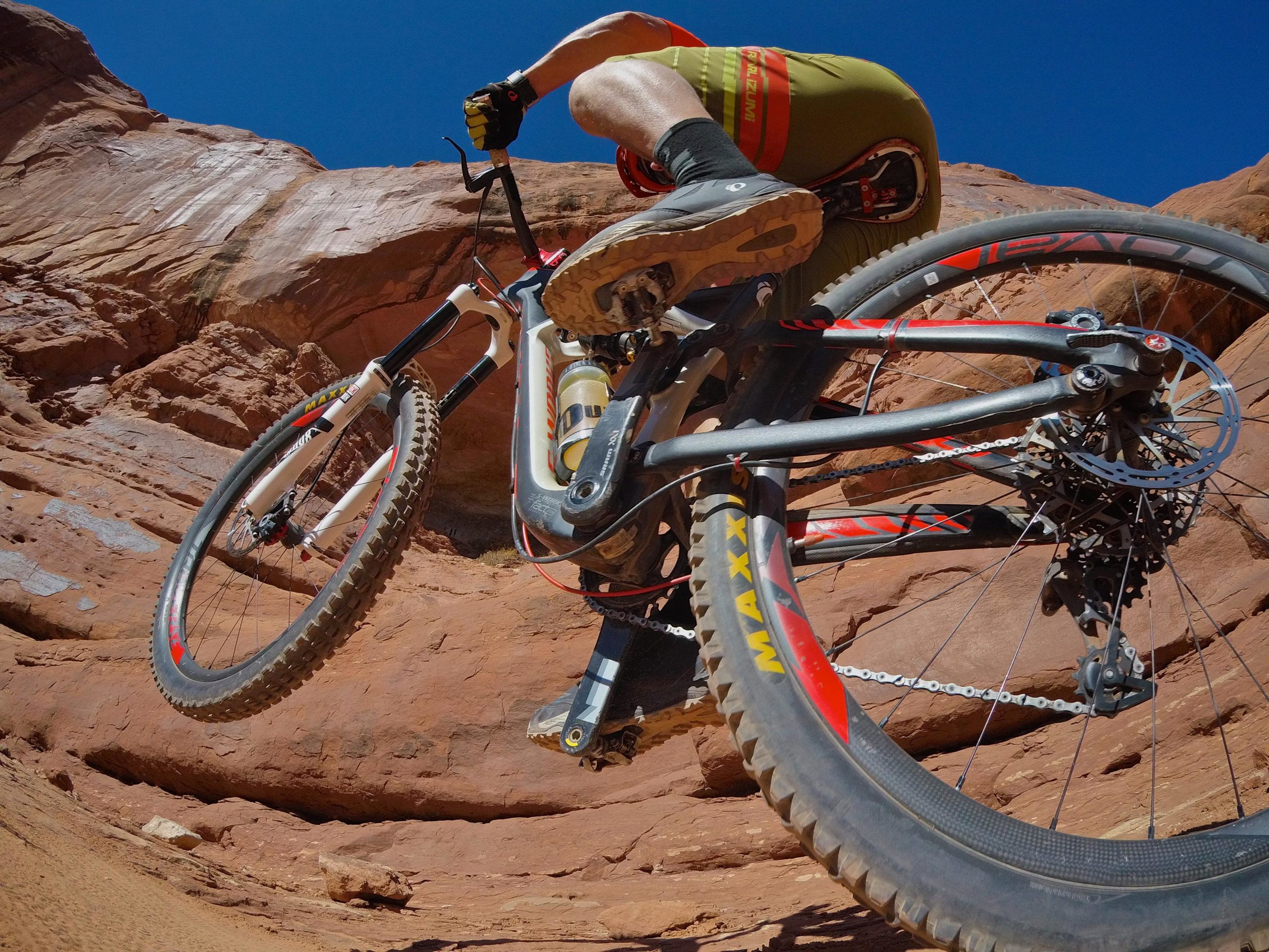 A mountain biker performing a jump on rocky terrain under a clear blue sky, showcasing the bike's wheels and a close-up of the rider's legs and bike frame. Big Mesa mountain bike trail.