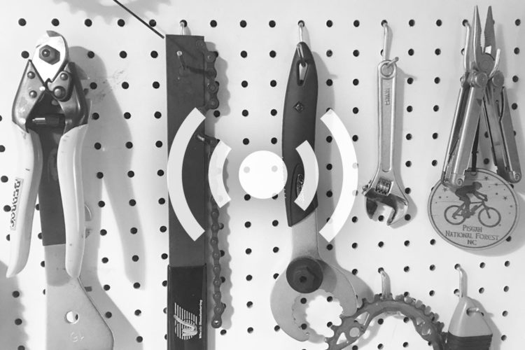 A collection of various bicycle tools hanging on a white pegboard, including a pair of wire cutters, a chain whip, a multi-tool, and an adjustable wrench, alongside a keychain featuring a bicycle silhouette and "Pisgah National Forest" engraved on it.