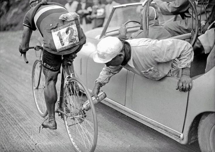 Specialized Camber Comp 29: A black and white photograph of a cyclist in a race wearing a numbered jersey, leaning towards a car where a crew member leans out of the window handing a water bottle to the cyclist. The scene captures the dynamic of support during a competitive cycling event, with spectators in the background observing the activity.
