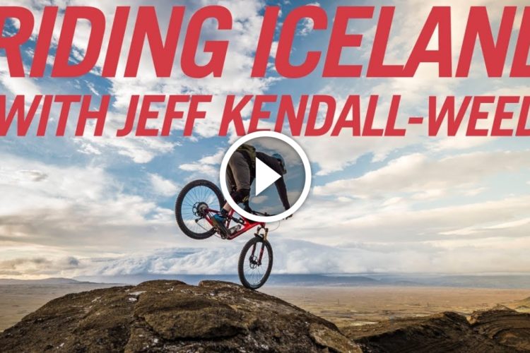 A mountain biker performing a stunt on a rocky outcrop with a scenic landscape of Iceland in the background. The image features bold text overlay that reads "Riding Iceland with Jeff Kendall-Weed."