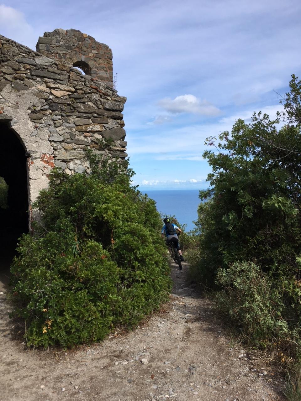 A cyclist riding a mountain bike along a path next to lush greenery and a stone ruin, with a view of the ocean in the background under a partly cloudy sky. 24h finale into DH Donne mountain bike trail.