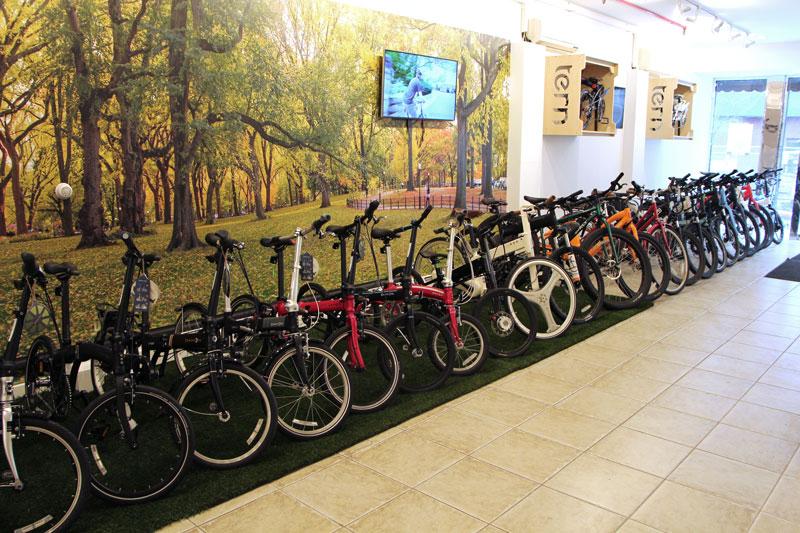 A display of various bicycles lined up indoors, featuring a mix of colors and styles. The background includes a vibrant mural of autumn trees, and a television screen is mounted on the wall, showing a cycling scene. The floor is tiled, with a patch of artificial grass beneath the bicycles, creating a welcoming atmosphere for customers.