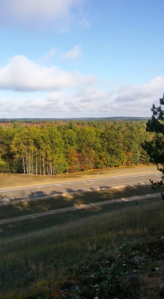 A scenic view of a rural highway bordered by a lush forest featuring a mix of green and autumn-colored trees under a partly cloudy sky. The foreground includes grassy terrain leading up to the road, with shadows cast by trees on the pavement. Hanson Hills mountain bike trail.