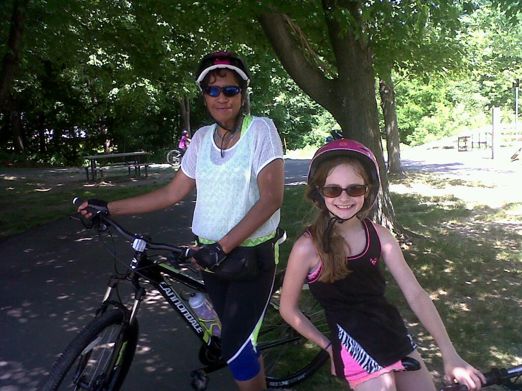 Cannondale Trail SL 29 2: Two cyclists pose for a photo on a sunny day. One adult, dressed in a white mesh top and bike shorts, stands next to a mountain bike, while a young girl in a black tank top and a pink helmet smiles brightly while sitting on her bike. They are surrounded by greenery, with trees and park amenities visible in the background.