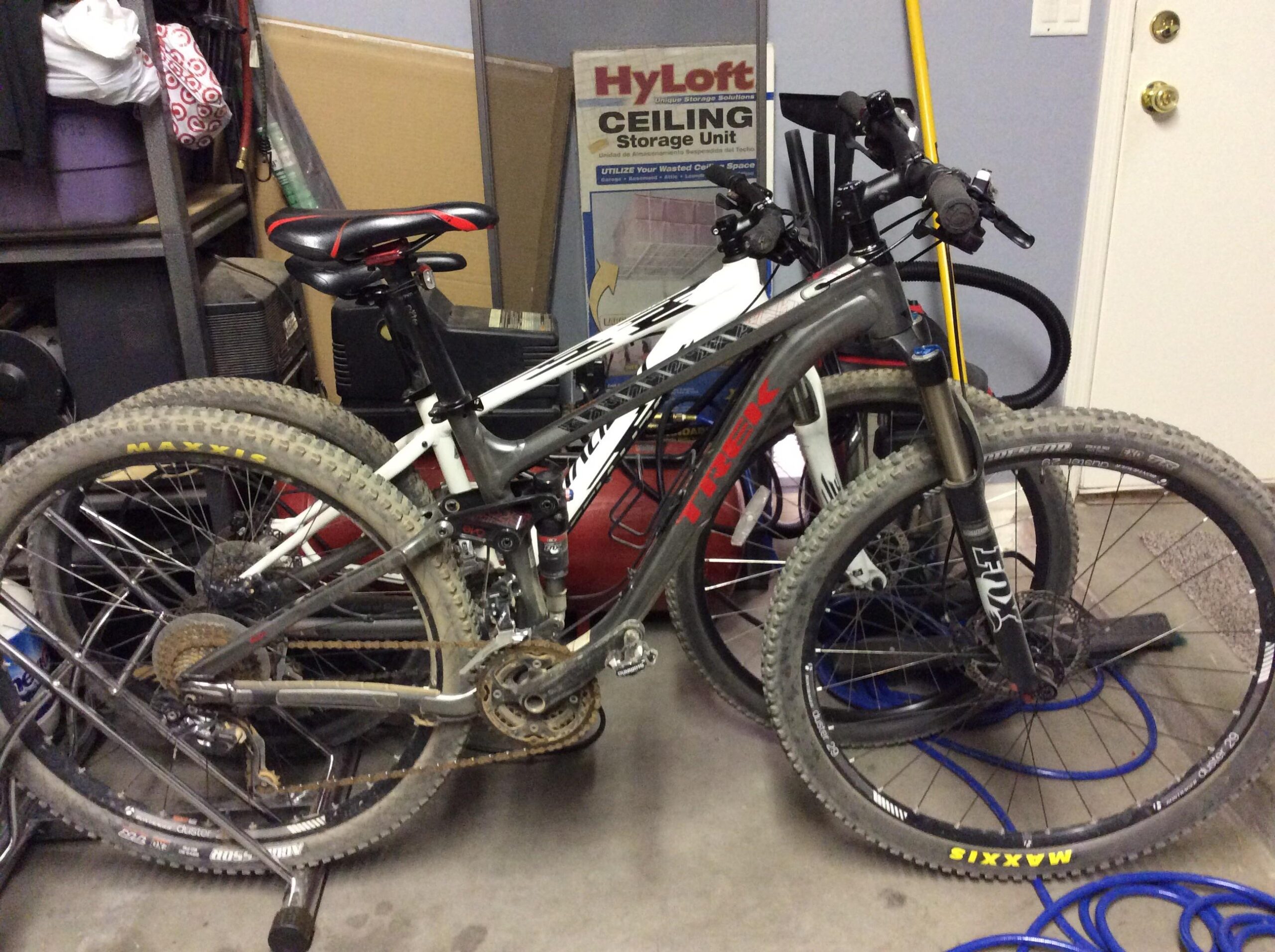 Trek Fuel EX 8: Two mountain bikes are parked side by side in a garage. The bike on the left is predominantly black and gray, featuring a red accent, and shows signs of use with dirt on the tires. The bike on the right is mostly white and appears to be in a similar condition. Background elements include shelves with storage bins and a door leading outside.