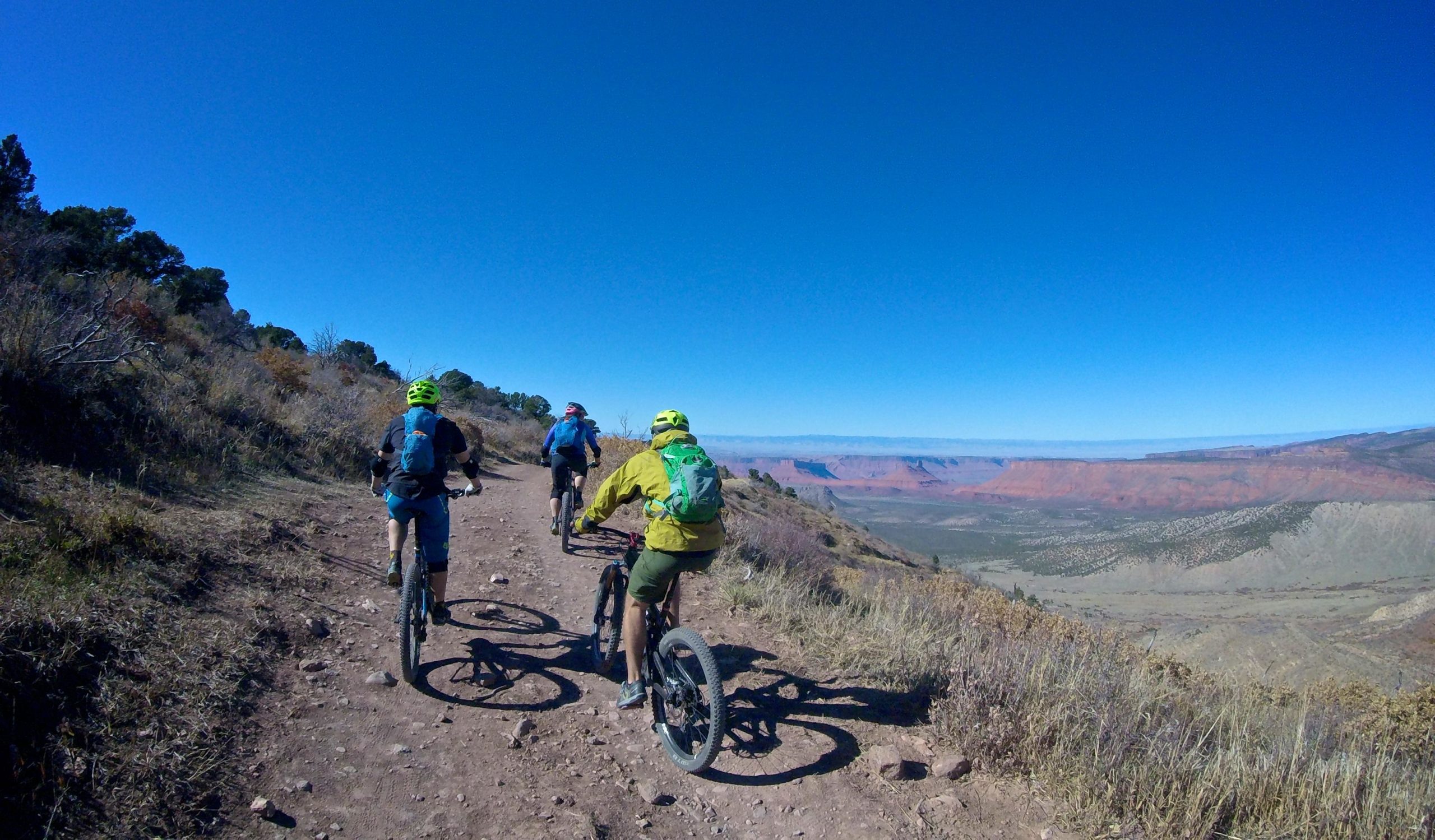 Four mountain bikers ride along a dirt trail with a panoramic view of a colorful landscape featuring red rock formations and rolling hills under a clear blue sky. The bikers are wearing helmets and backpacks, with the trail surrounded by sparse vegetation and brush. The Whole Enchilada mountain bike trail.