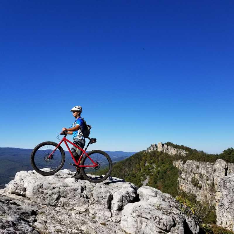 A person standing on a large rock outcrop, holding a red mountain bike, with a clear blue sky and mountains in the background. The individual is wearing a helmet and casual biking attire. North Fork Mountain Trail mountain bike trail.