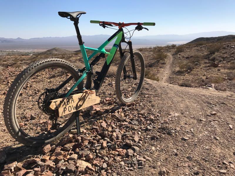 A mountain bike is parked on a rocky trail with a wooden sign labeled "TAMBA POW." The backdrop features rolling hills and a clear blue sky, suggesting a bright, sunny day suitable for outdoor activities. Bootleg Canyon mountain bike trail.