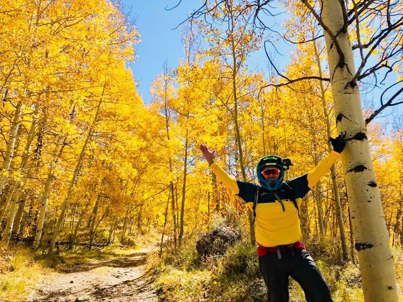 A person wearing a helmet and outdoor gear stands on a trail surrounded by vibrant yellow aspen trees under a clear blue sky, raising their arms in celebration. Strand Hill mountain bike trail.