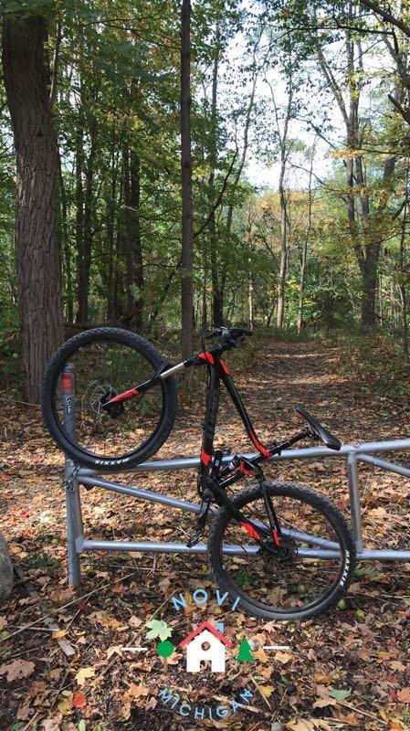 A mountain bike resting on a silver railing in a wooded area during autumn. The ground is covered with fallen leaves in various shades of brown and yellow. In the background, tall trees display a mix of green and golden foliage. A graphic at the bottom reads "Novi, Michigan," featuring a small house and trees. Novi Tree Farm (Lakeshore Park) mountain bike trail.