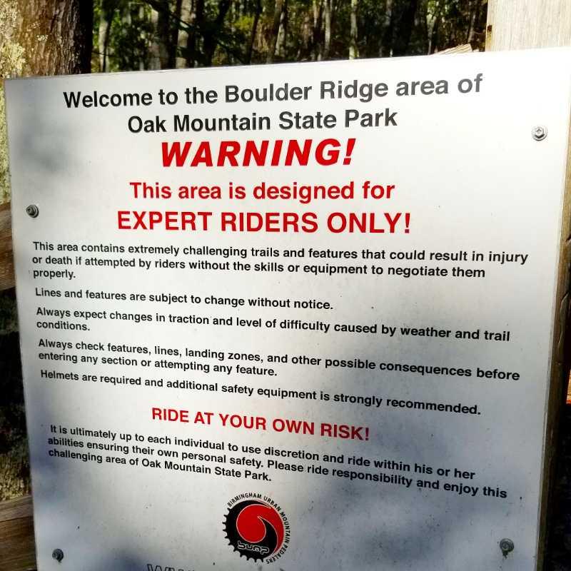 Sign at Oak Mountain State Park warning that the Boulder Ridge area is for expert riders only. It emphasizes the challenging trails and features that could lead to injury or death for inexperienced riders. The sign advises riders to expect changes in trail conditions, check features and landing zones before entering, and wear helmets along with additional safety gear. It concludes with a reminder to ride at one's own risk. Oak Mountain State Park mountain bike trail.