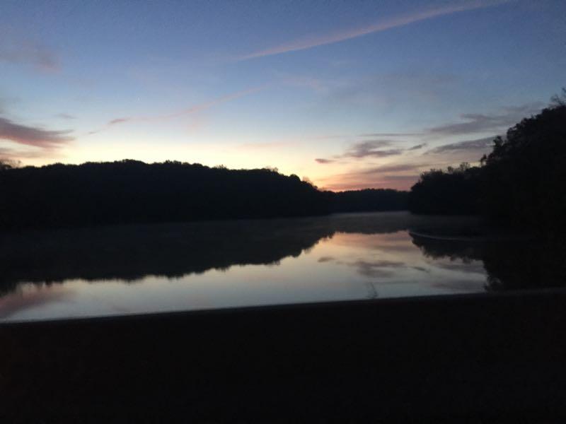 Scenic view of a tranquil river at dawn, with the silhouette of trees in the foreground and a soft gradient of colors in the sky transitioning from deep blue to warm orange and pink. The calm water reflects the colors of the sky, creating a serene atmosphere. Fountainhead Regional Park mountain bike trail.