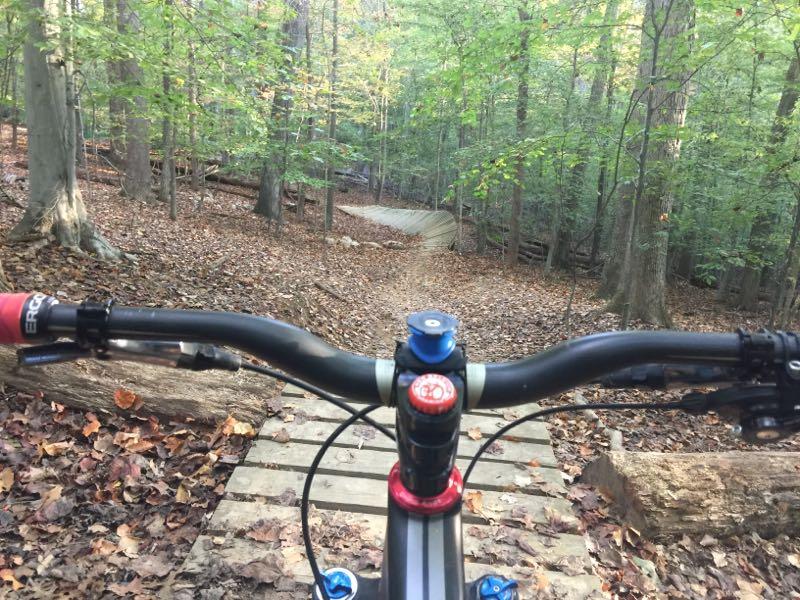 View from the handlebars of a mountain bike, looking down a forested trail with wooden planks and scattered leaves. The scene features tall trees with green foliage and a winding path ahead, indicating a downhill descent in a natural setting. Fountainhead Regional Park mountain bike trail.