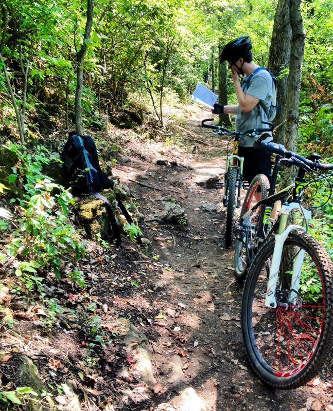 A person in a grey shirt and black helmet stands on a dirt trail surrounded by lush greenery, holding a map or guide while looking thoughtful. Two mountain bikes are parked nearby, and a backpack rests against a rock. The scene captures a moment of exploration in a natural setting. Castlewood State Park mountain bike trail.