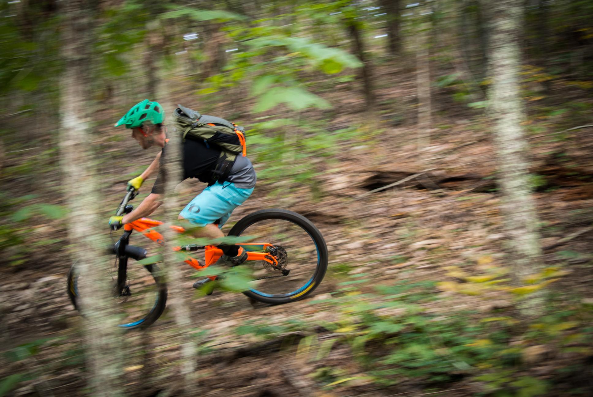 Orange Stage 5 RS: A cyclist riding a mountain bike quickly down a wooded trail, surrounded by trees and foliage. The cyclist is wearing a green helmet, a black shirt, and blue shorts, and is carrying a backpack. The motion blur conveys speed and excitement.