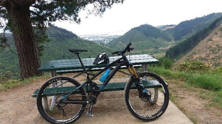 Mondraker Foxy XR: A mountain bike parked next to a picnic table with a scenic view of green hills and a valley in the background. A black helmet is placed on the bike, and there are trees surrounding the area. The sky is overcast.