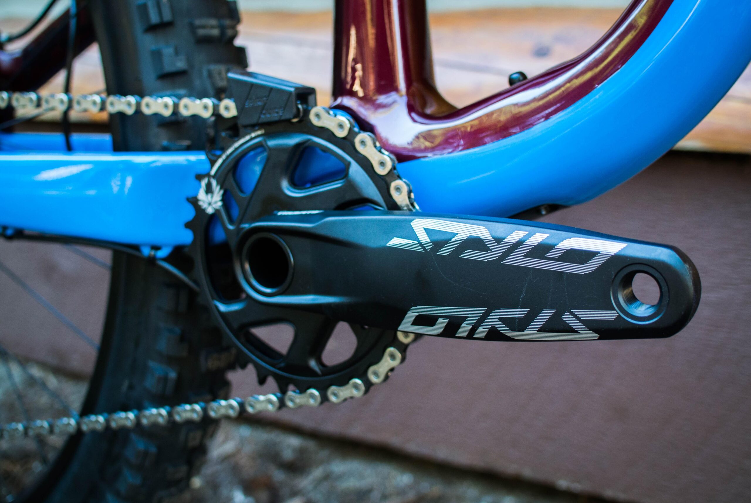 Rocky Mountain Pipeline: Close-up view of a mountain bike's crankset, featuring a black crank arm with the logo "SNYLO," a chainring, and a portion of the bike's blue frame. The background shows part of a brown wooden surface and the tread of the bike tire.