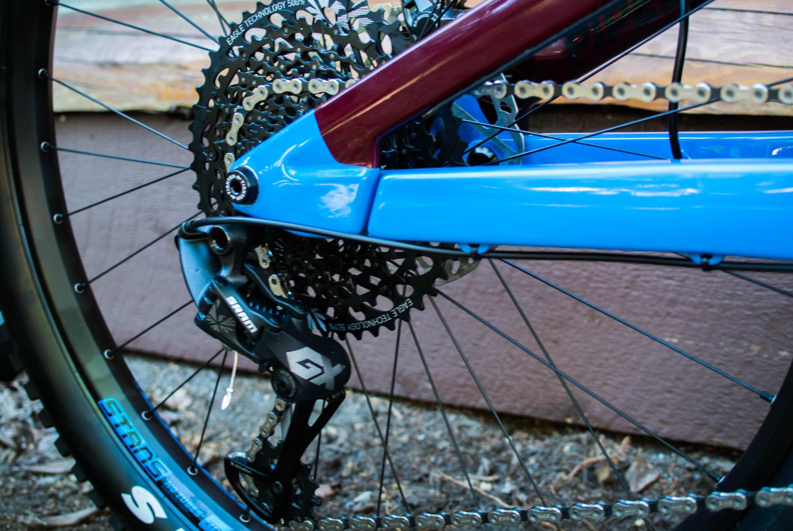 Rocky Mountain Pipeline: Close-up of a mountain bike's rear drivetrain, showcasing the blue and maroon frame, derailleur, and cassette. The background features a wooden structure and gravel.