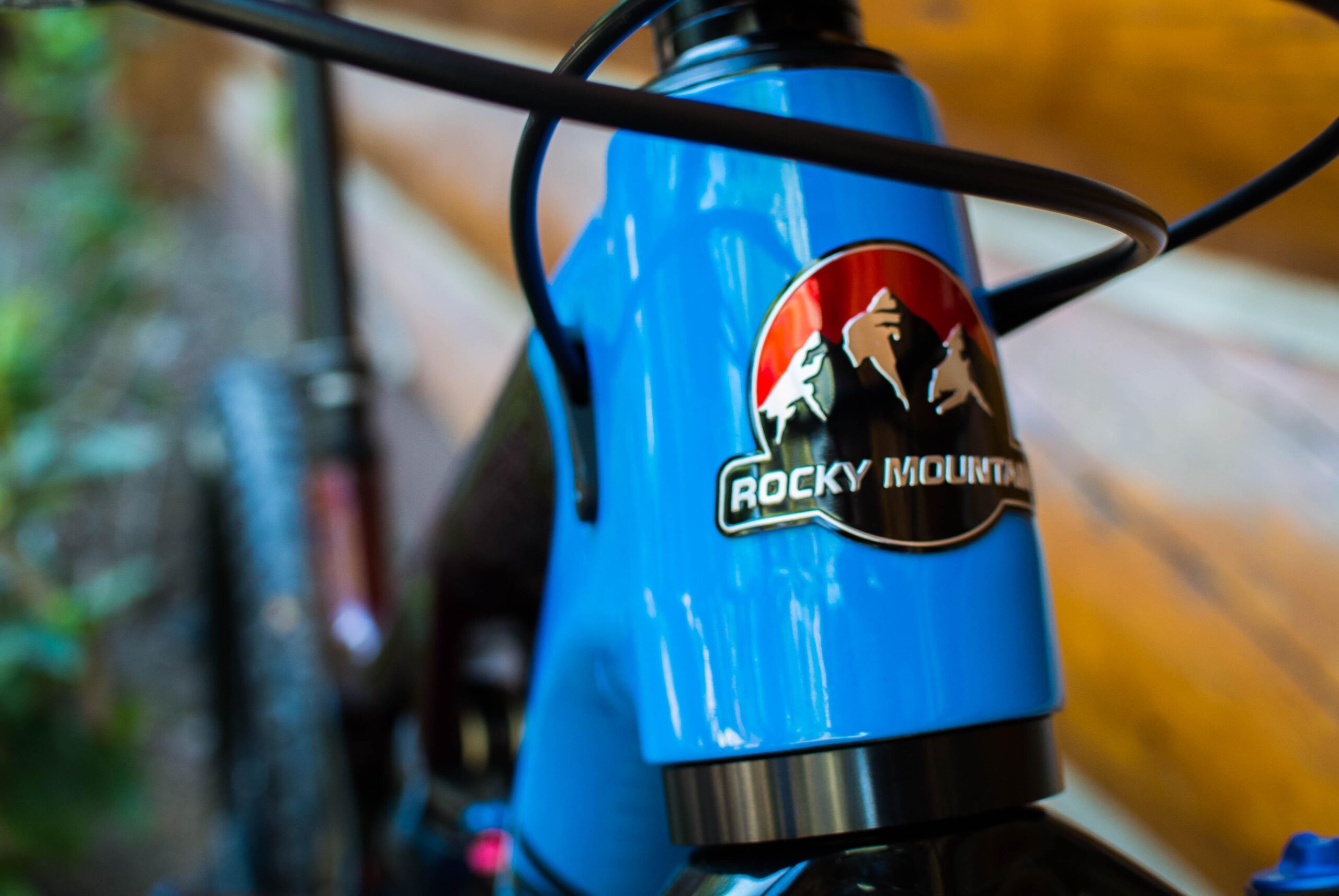 Rocky Mountain Pipeline: Close-up of a blue mountain bike frame featuring the "Rocky Mountain" logo prominently displayed on the head tube, with blurred natural elements and wooden background enhancing the outdoor setting.