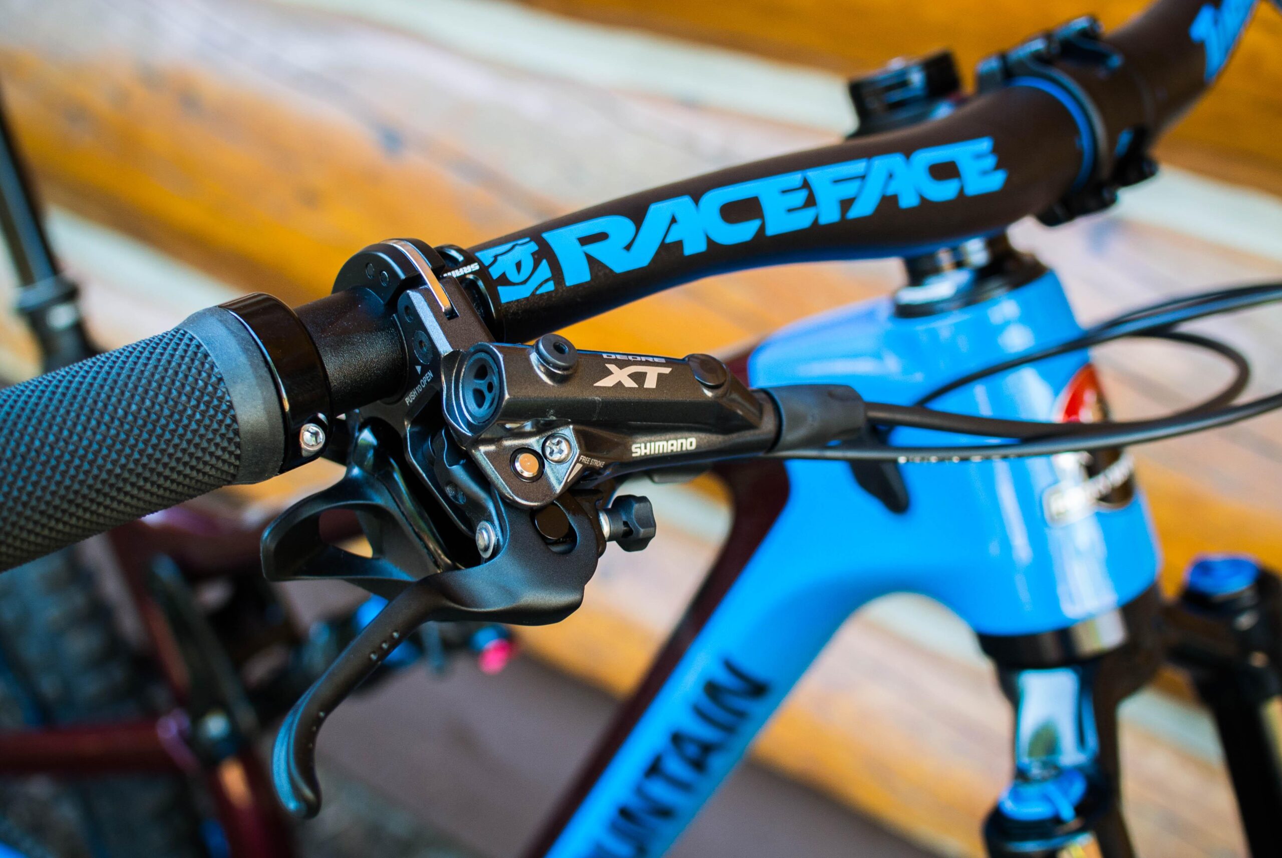 Rocky Mountain Pipeline: Close-up view of a mountain bike handlebar featuring a Shimano XT brake lever and a textured grip. The handlebar is branded with "RaceFace" and the bike frame is blue with a visible "Mountain" inscription. A wooden background is partially visible.
