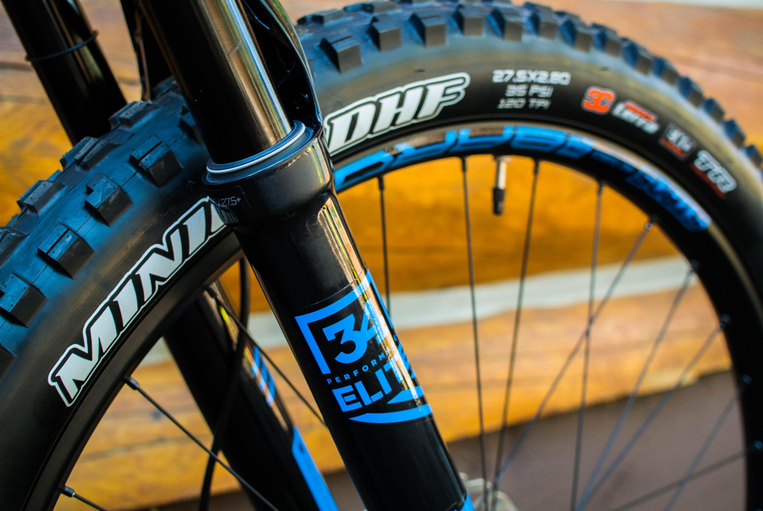 Rocky Mountain Pipeline: Close-up of a mountain bike's front fork and tire, featuring a black and blue color scheme. The tire has a tread pattern designed for traction, with visible branding details on the fork and tire sidewall. The background is blurred wooden planks.