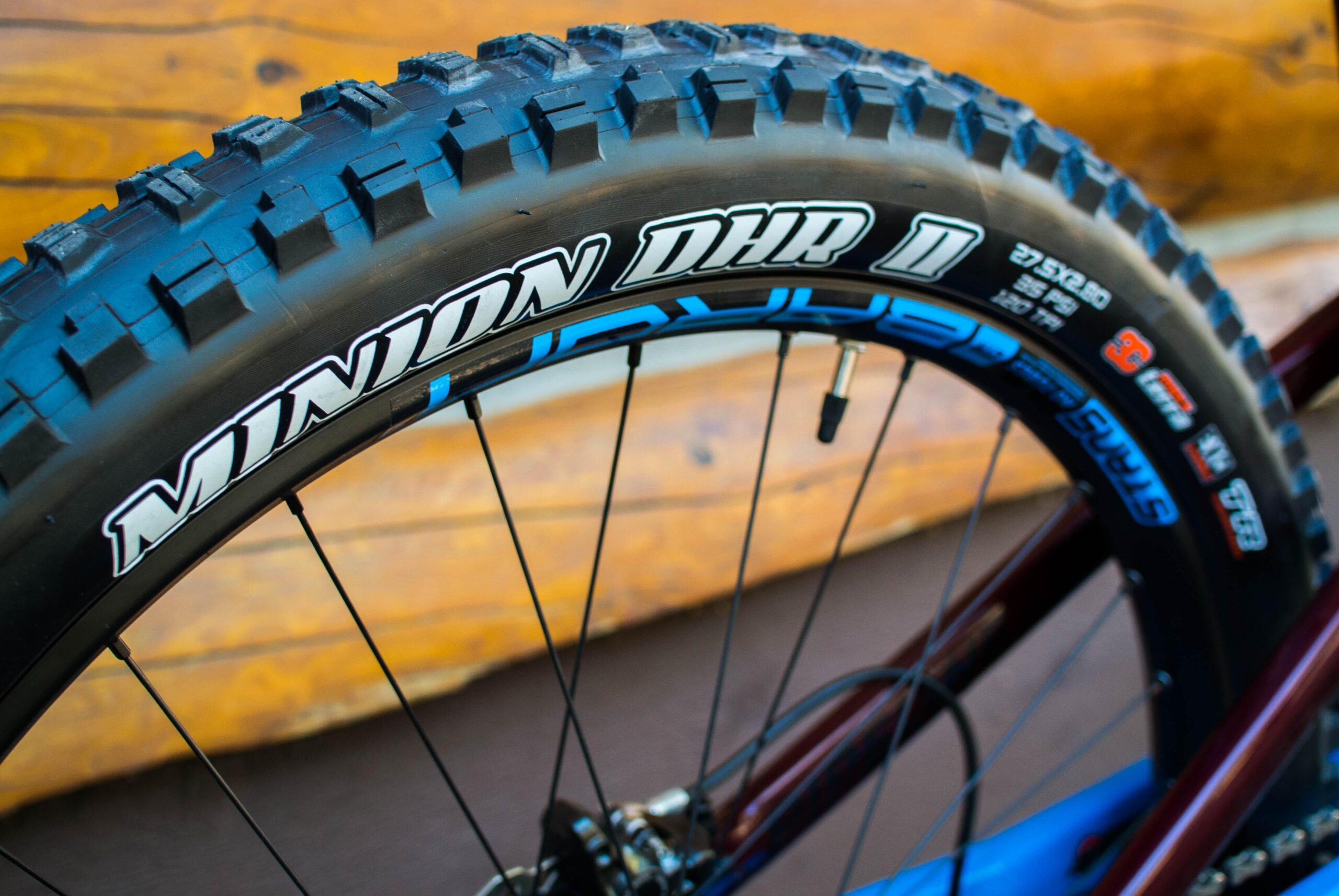 Rocky Mountain Pipeline: Close-up of a mountain bike tire featuring a heavily treaded design, identified as a Minion DHR II, mounted on a dark rim. The background showcases a wooden surface, providing a natural outdoor context.