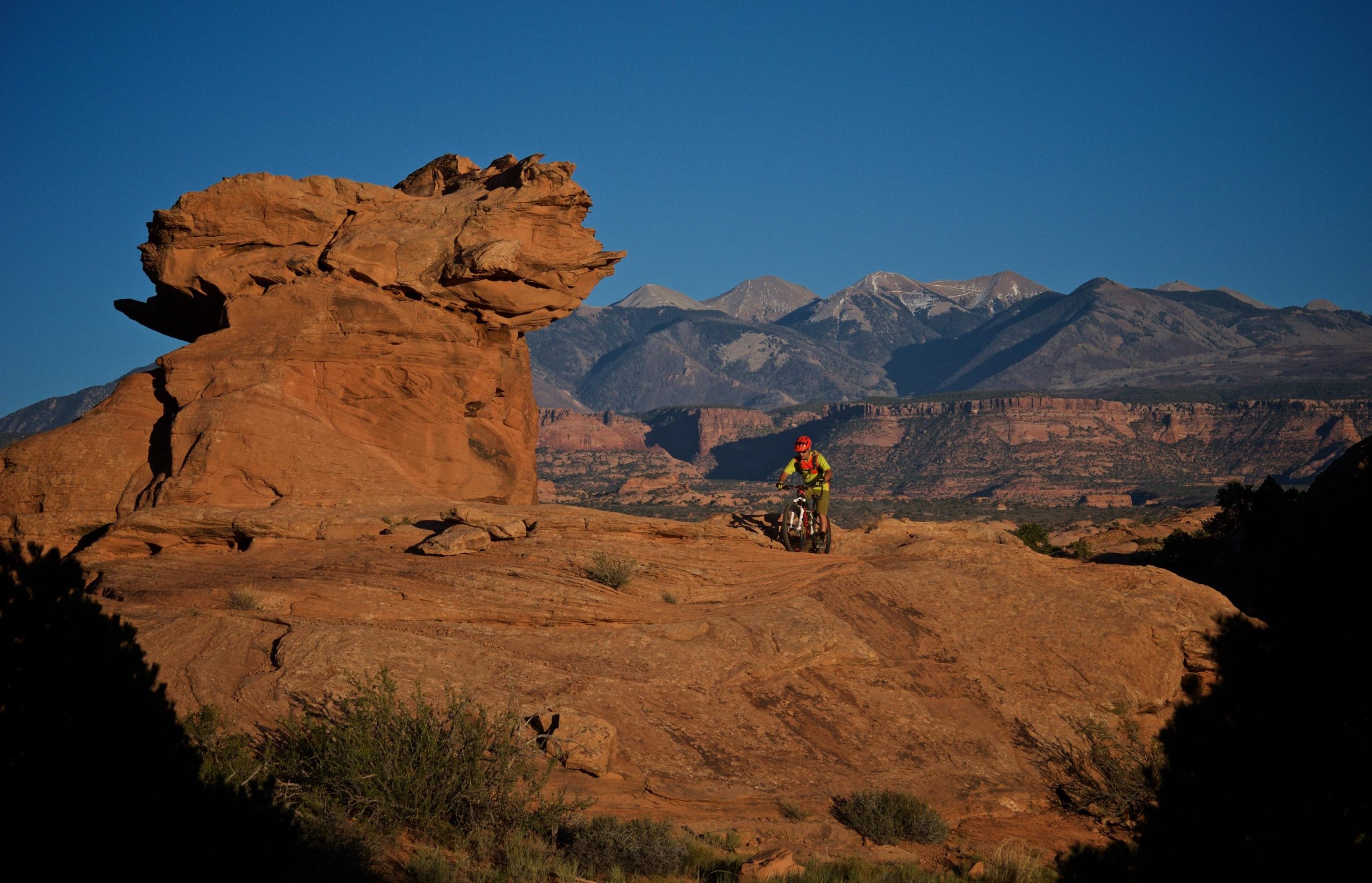 A mountain biker navigates rocky terrain with a large, uniquely shaped rock formation in the foreground and snow-capped mountains visible in the background under a clear blue sky. Slickrock mountain bike trail.