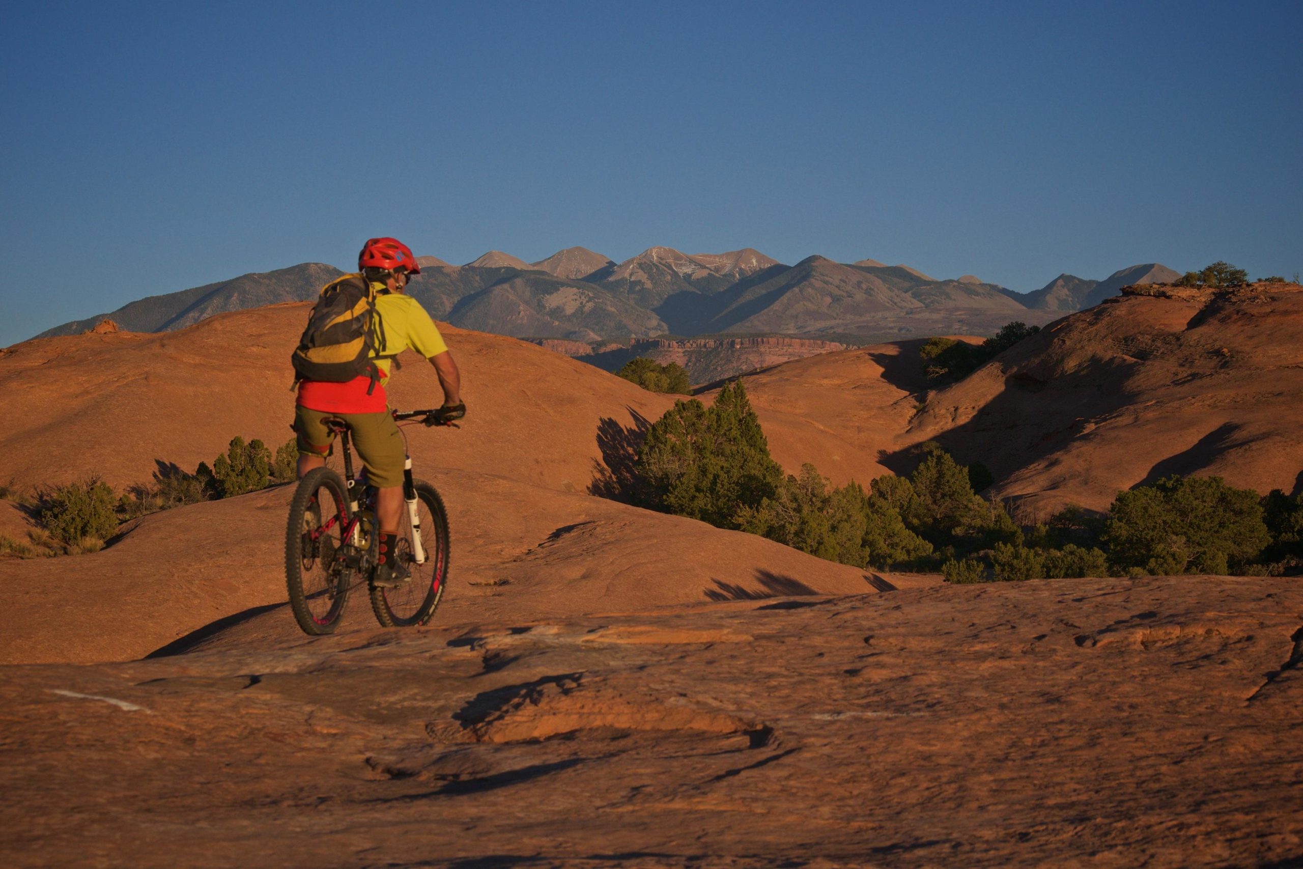 A mountain biker wearing a bright yellow shirt and red helmet is riding on rocky terrain, overlooking a scenic landscape of rolling hills and distant mountains under a clear blue sky. The sunlight casts warm tones over the rocky surface, highlighting the natural beauty of the outdoor setting. Slickrock mountain bike trail.