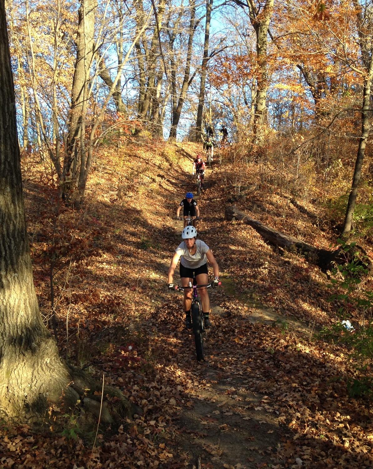 A mountain biker navigating a leaf-covered trail in a wooded area during autumn. The path slopes upward, with several other cyclists visible in the background. Trees with autumn foliage line the trail under a clear blue sky. Sunbridge Trail mountain bike trail.
