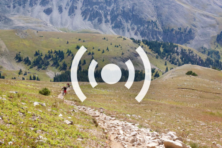 A scenic mountain landscape featuring hikers on a rocky trail surrounded by lush green grass and trees, with a backdrop of towering mountains under a clear blue sky. A stylized sound icon is overlaid on the image.