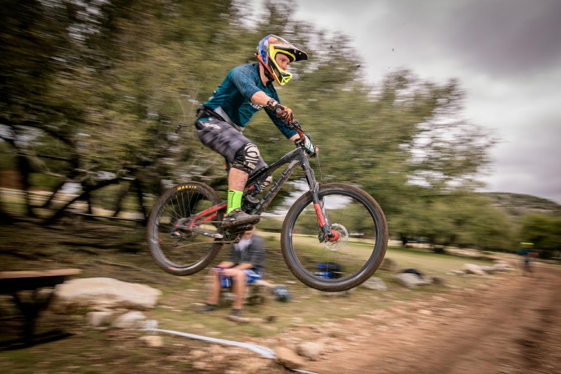 Intense Tracer 275c: A mountain biker in a blue shirt and helmet performs a jump over a dirt path, with trees in the background. The bike is mid-air, showcasing the rider's skill as they navigate a trail. In the background, a few spectators can be seen watching the jump.