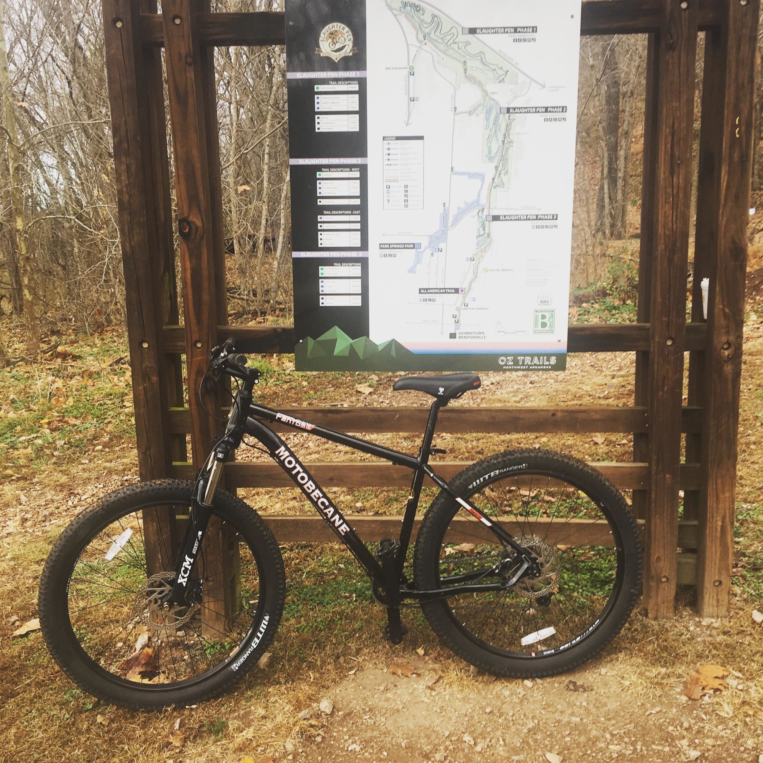 A black mountain bike parked next to a wooden trail sign displaying a map of the Oz Trails. The background features bare trees and a leaf-covered ground, indicating a fall or early winter setting. Slaughter Pen Trail mountain bike trail.