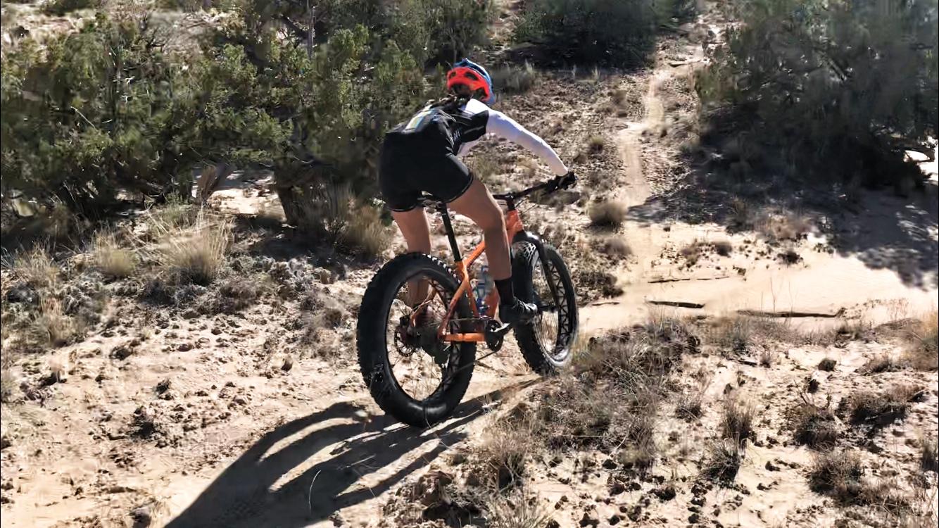 A cyclist riding a fat bike on a sandy, uneven trail surrounded by sparse desert vegetation. The rider wears a bright helmet and athletic gear, showcasing an action shot as they navigate the rugged terrain. Mariposa Fat Bike Trails mountain bike trail.