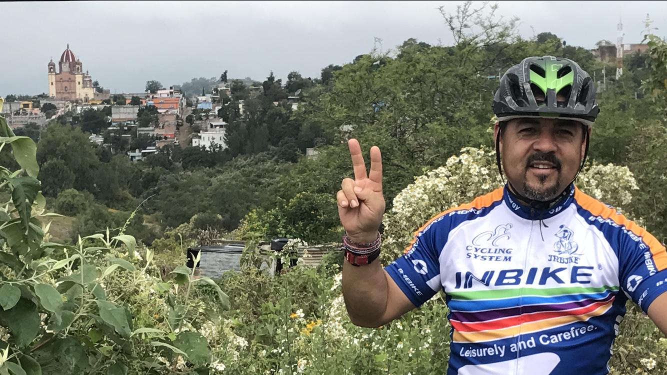 Cannondale F29 6: A cyclist in a colorful jersey poses with a peace sign against a scenic backdrop of hills and a town, featuring a church with a distinctive dome. Lush greenery and wildflowers surround the cyclist in the foreground, while the town with colorful buildings is visible in the distance under a cloudy sky.
