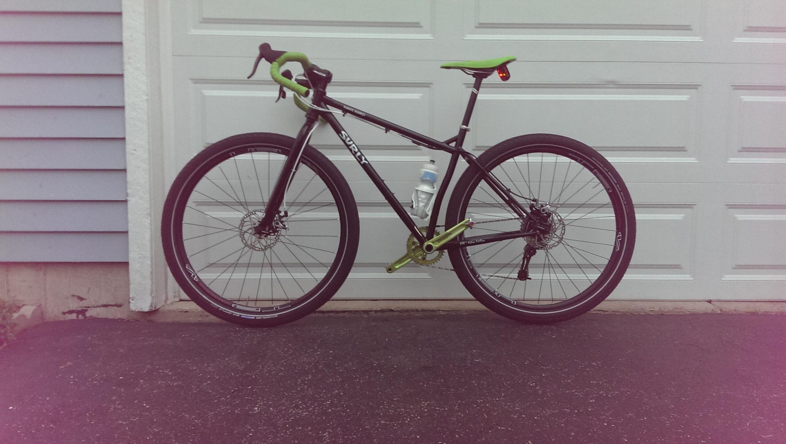 Surly Karate Monkey: A black Surly bicycle with green accents, featuring a water bottle attached, parked on asphalt in front of a white garage door. The bike is equipped with disc brakes and has road-style handlebars.