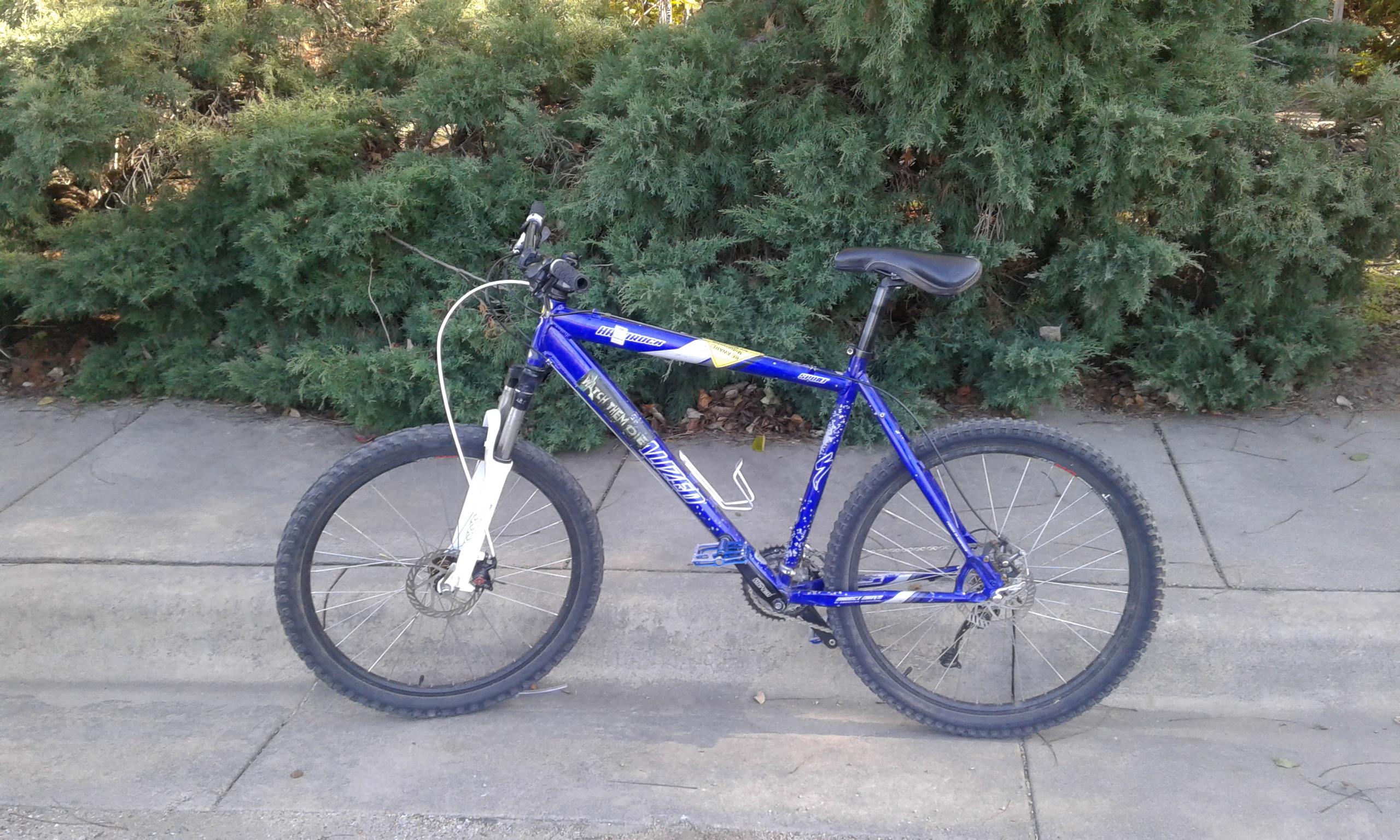 Specialized hardrock: A blue mountain bike parked beside a hedge, on a concrete sidewalk. The bike features thick tires and a white front fork, with visible gear and brake components.