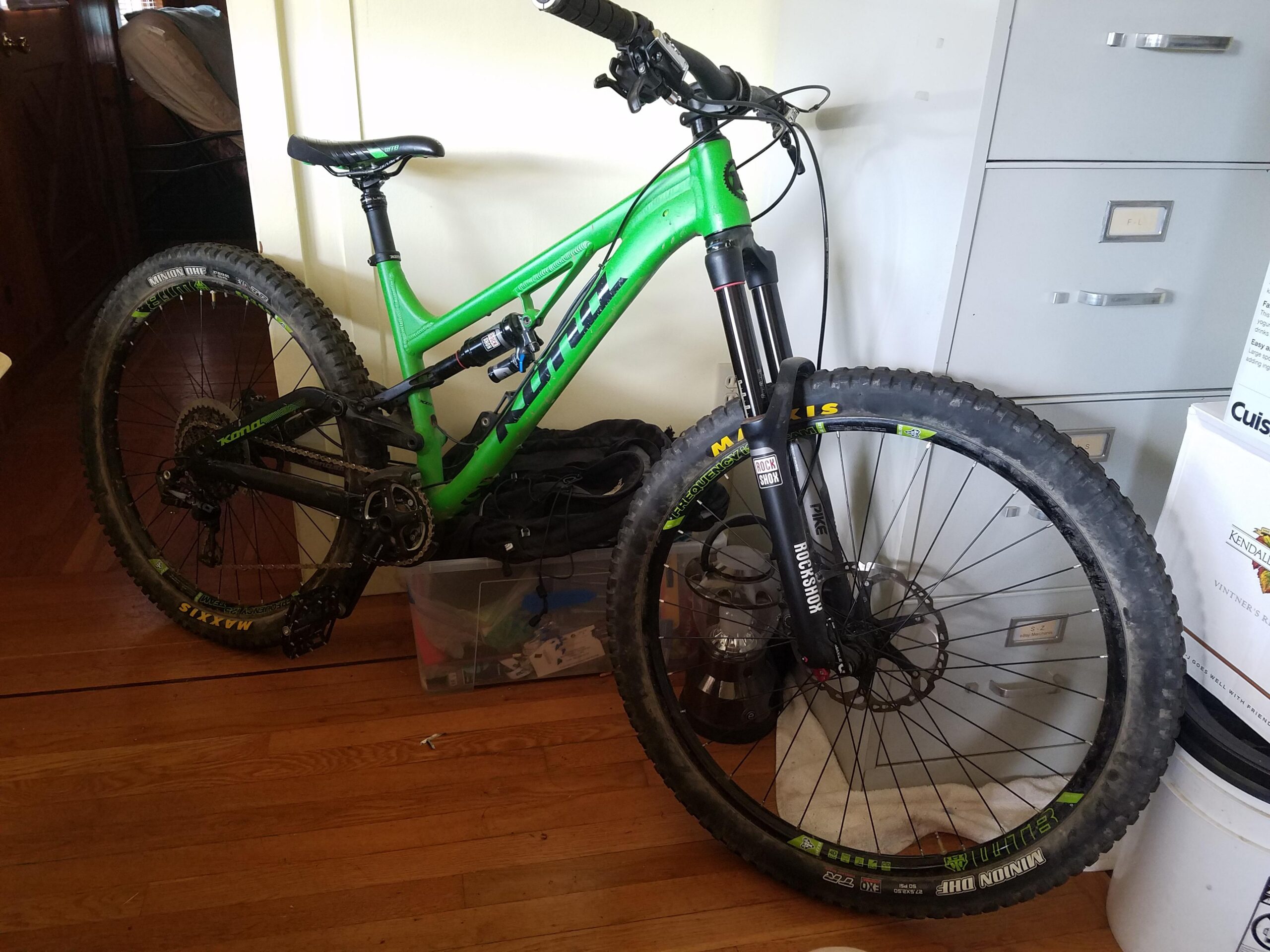 Kona Process 153 DL: A green mountain bike leaning against a wall, featuring thick tires and a suspension fork. The bike is positioned near a filing cabinet and other items, with wooden flooring visible in the foreground.