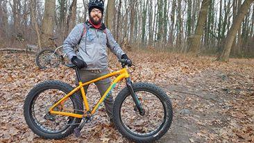 Specialized Fatboy: A person wearing a helmet and outdoor gear stands next to a large fat bike on a forest trail covered with fallen leaves. Trees are visible in the background, indicating a natural setting.