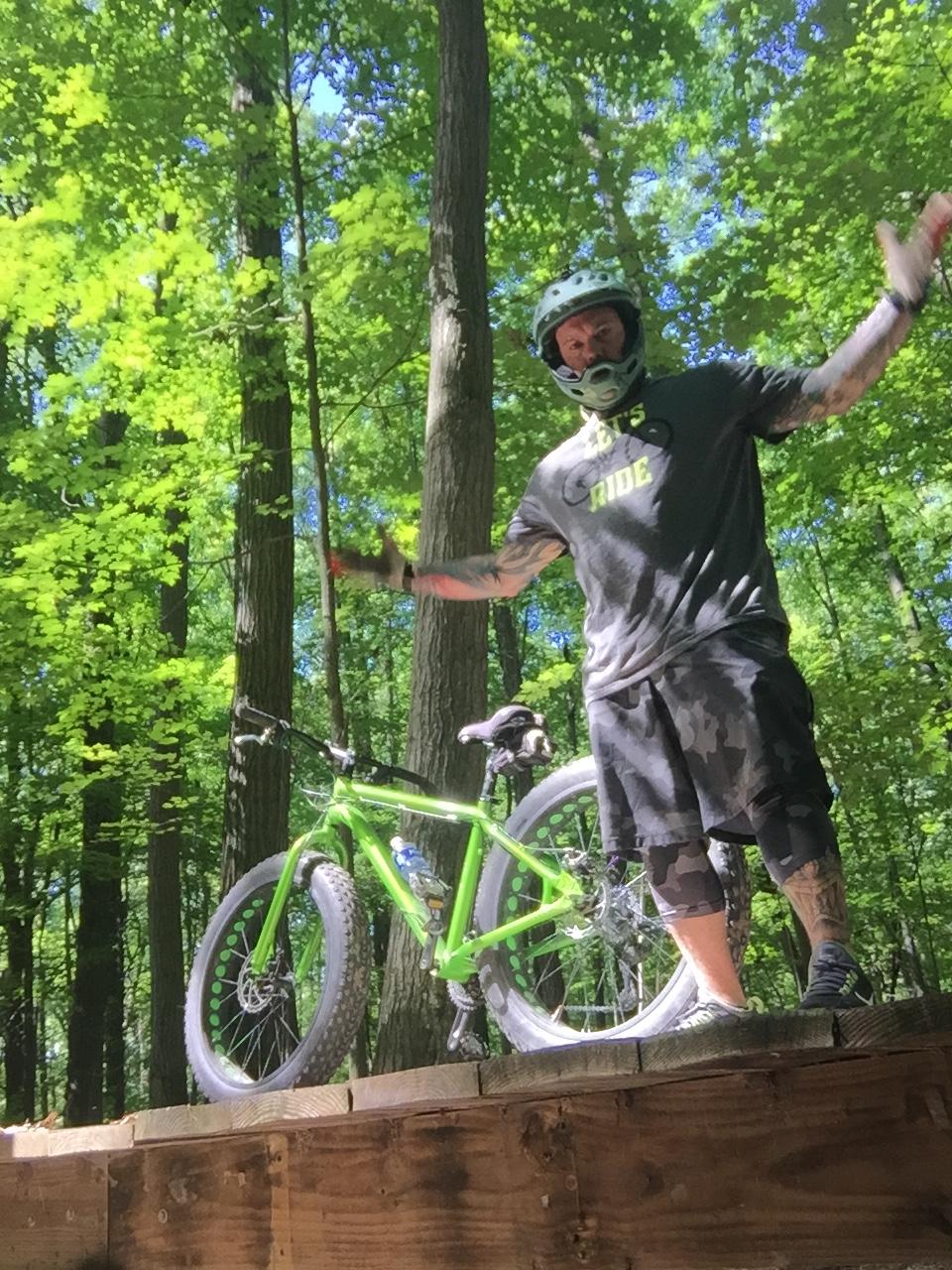 Giant Stance 27.5 2: A person in a helmet and casual athletic wear stands on a wooden ramp in a forested area, gesturing with their arms. A green fat bike is positioned beside them on the ramp, and lush green trees surround the scene, indicating a sunny day.