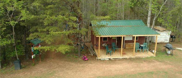 A small cabin surrounded by trees, featuring a wooden porch with green chairs, a green metal roof, and a grill beside it. A signpost is visible nearby, along with a dirt path leading to the cabin. Mountain Laurel Trails mountain bike trail.