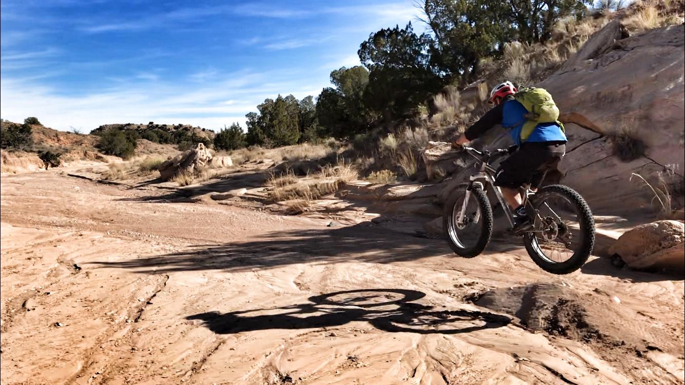 A mountain biker in a blue shirt and black shorts is riding a fat-tire bike over a sandy, rocky terrain under a clear blue sky. The rider is mid-jump, casting a shadow on the ground as they navigate the uneven landscape surrounded by shrubs and trees. Mariposa Fat Bike Trails mountain bike trail.