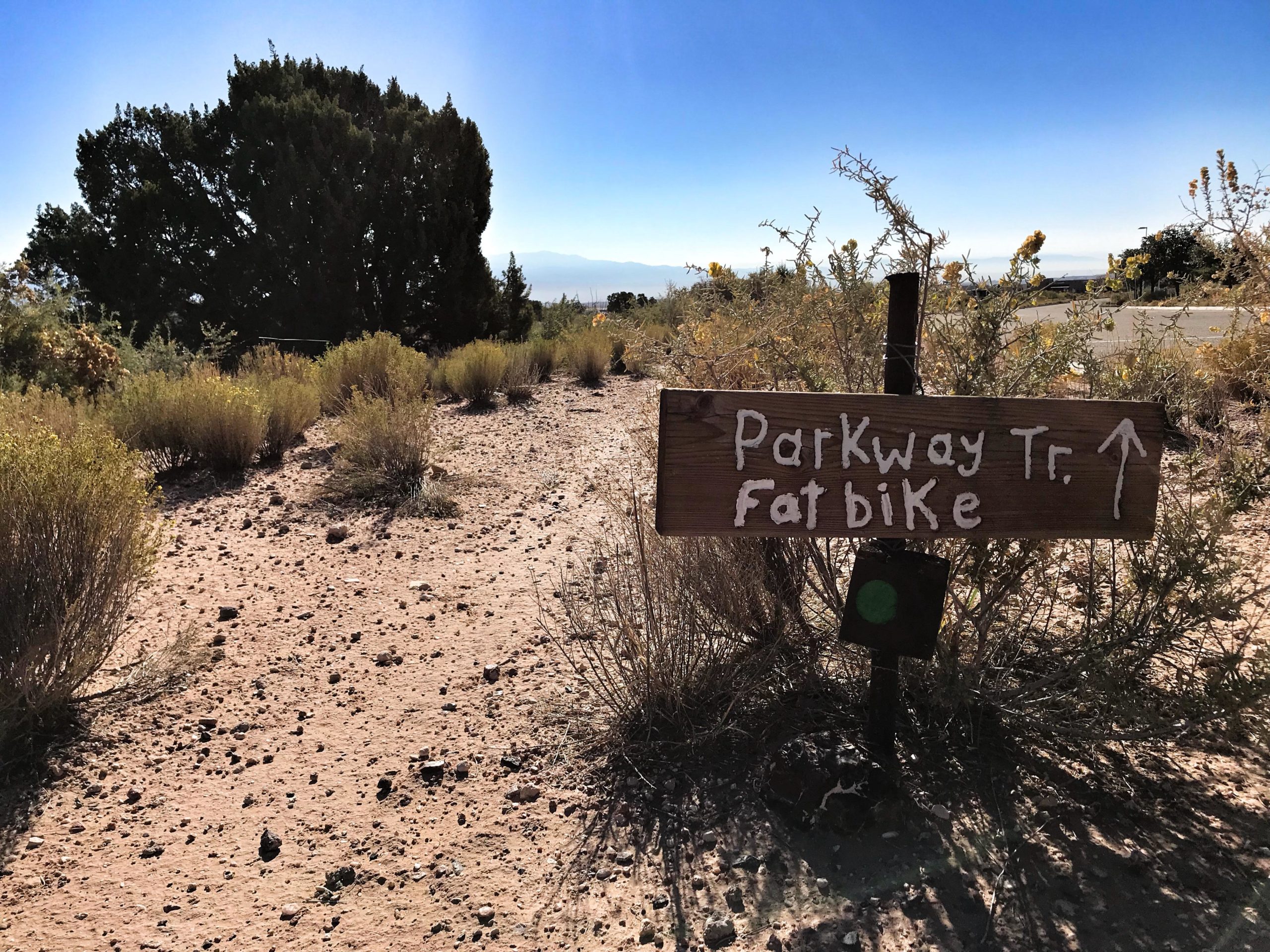 Signpost indicating the direction to "Parkway Tr. Fatbike" with a brown wooden sign. Surrounding landscape features sparse vegetation, dirt pathway, and distant hills under a clear blue sky. Parkway Fatbike trail mountain bike trail.
