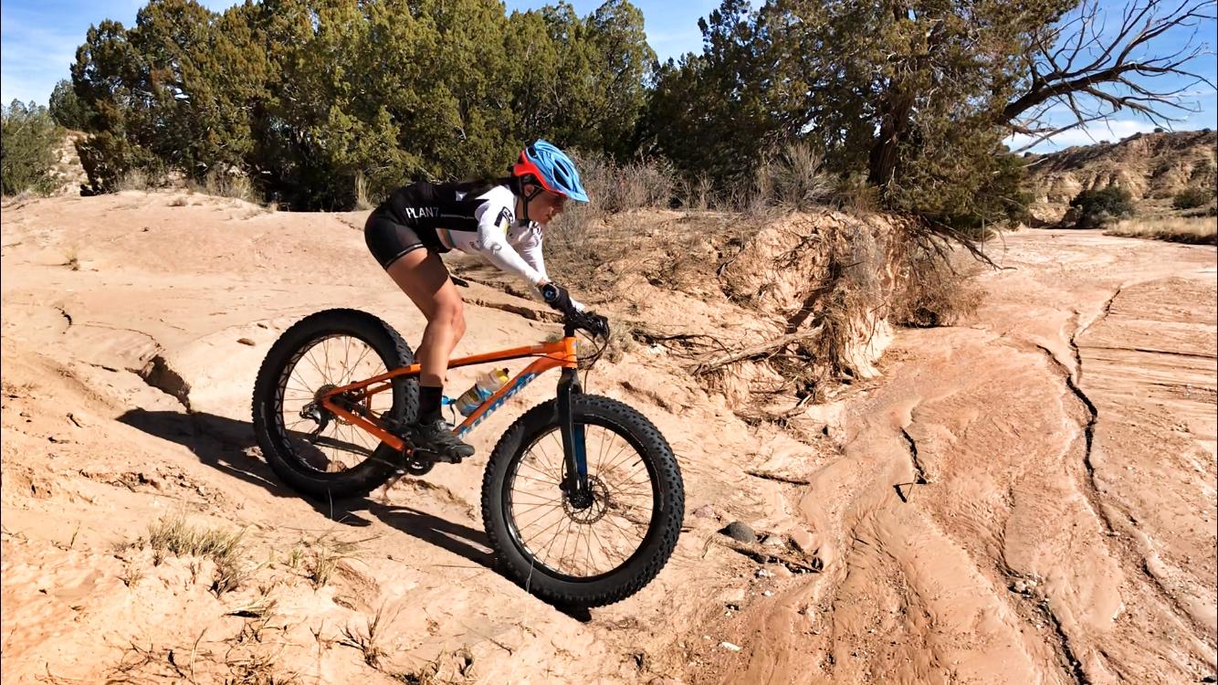 A female cyclist riding a bright orange fat bike on a rocky dirt trail, preparing to navigate an incline. She is wearing a blue helmet, a black and white cycling jersey, and black shorts. The background features sparse vegetation and a clear blue sky. Mariposa Fat Bike Trails mountain bike trail.