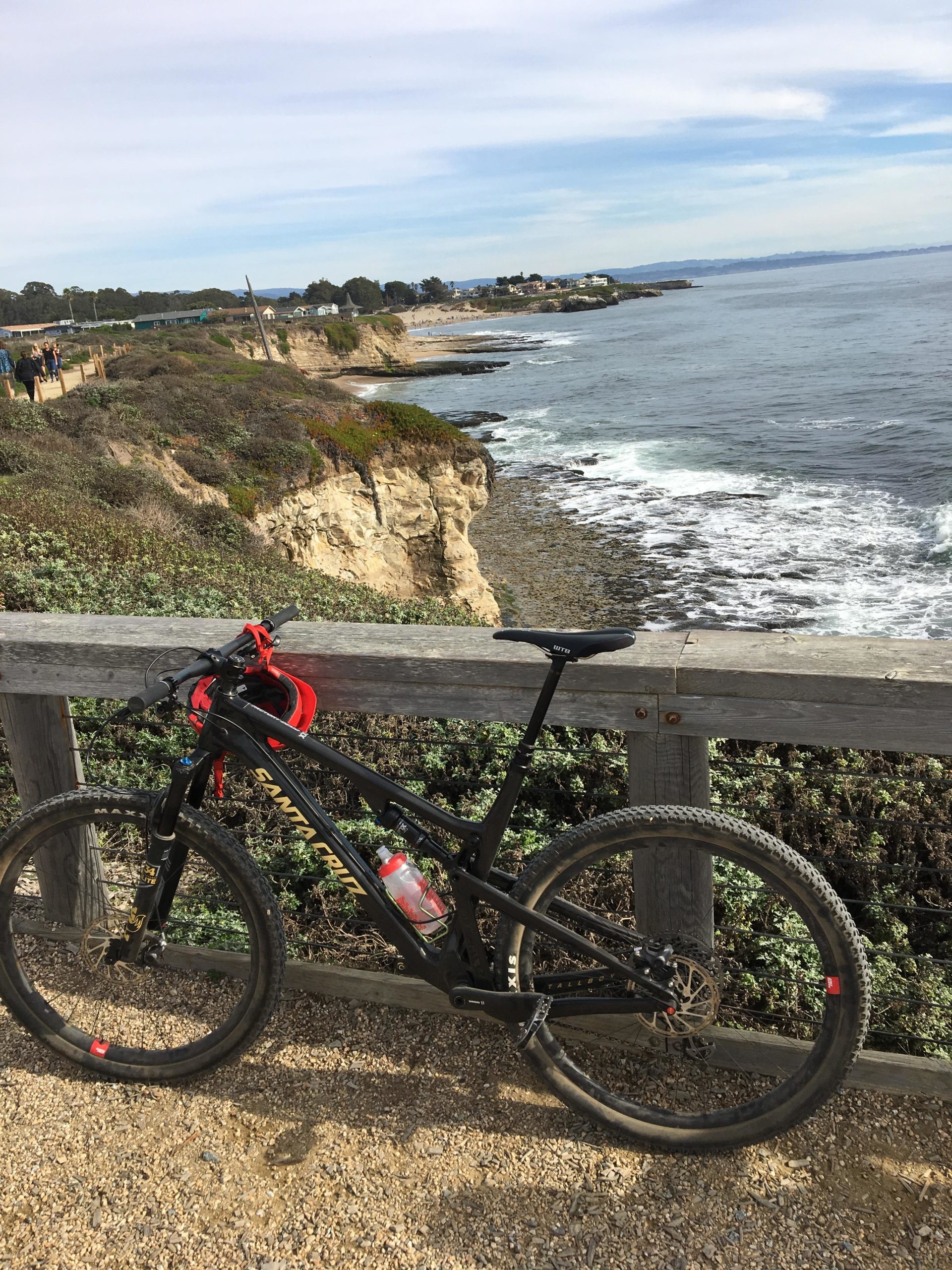 A mountain bike parked beside a wooden railing, overlooking a rocky coastline with waves crashing against the shore. In the background, a path is visible with people walking along the cliffside, and a small town is situated on the horizon under a blue sky with scattered clouds. Wilder Ranch State Park mountain bike trail.