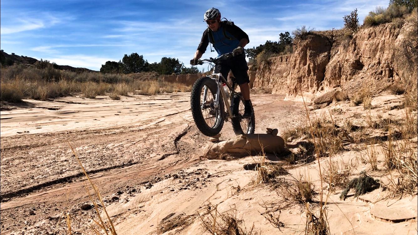 A person riding a mountain bike is performing a jump over a rock on a sandy trail surrounded by dry grass and rocky cliffs under a clear blue sky. Mariposa Fat Bike Trails mountain bike trail.