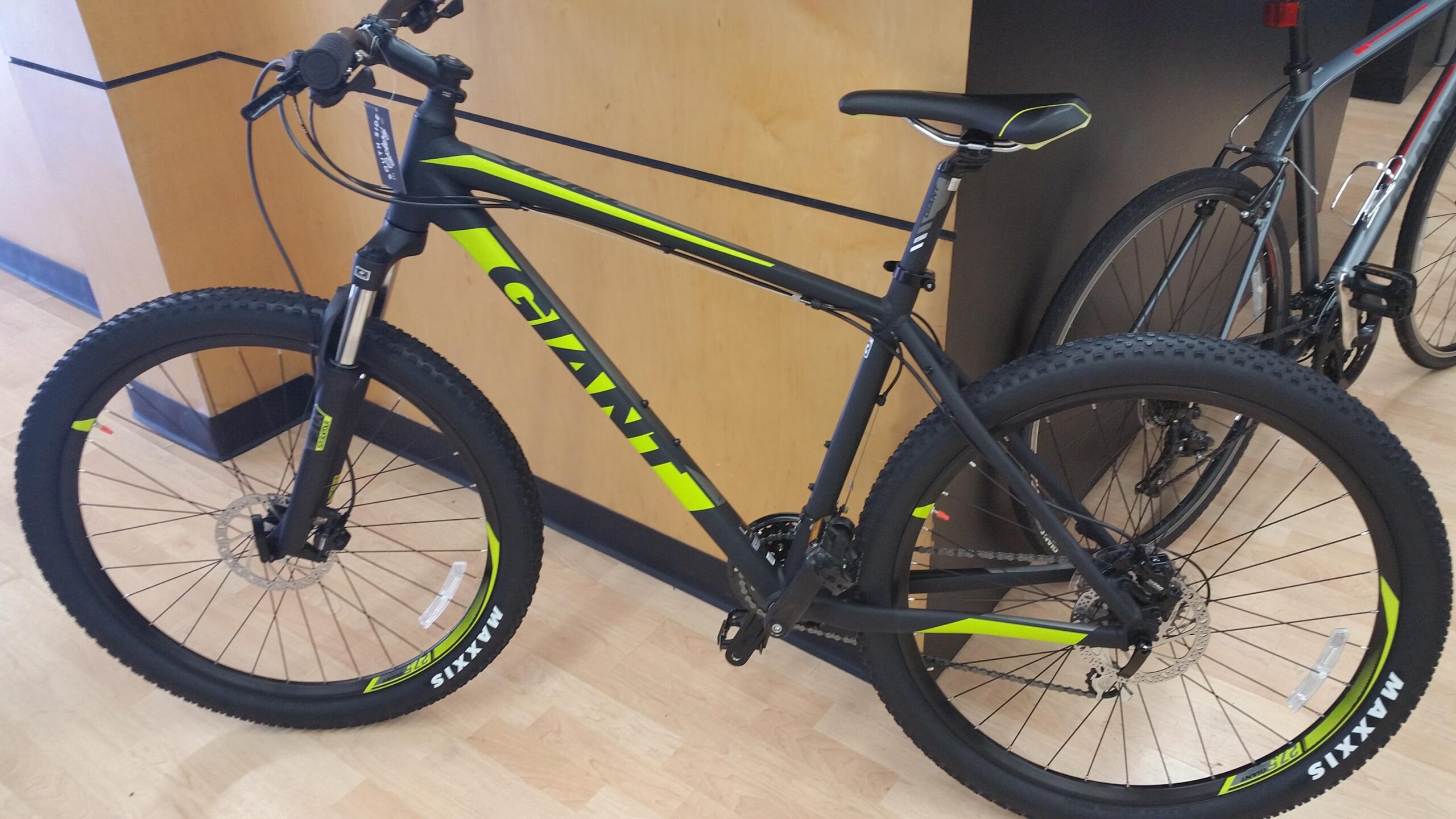 2017 Giant Talon 3: A black and neon green mountain bike displayed in a bicycle shop, featuring thick tires with Maxxis branding, a front suspension fork, and a sleek frame design. The backdrop includes a wooden wall and another bike partially visible.